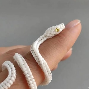 May include: A coiled snake ring crafted from small white beads, featuring a single yellow gemstone set as an eye. The flexible, woven beaded structure wraps around a finger, creating a textured, serpent-like appearance with a bright, singular accent.
