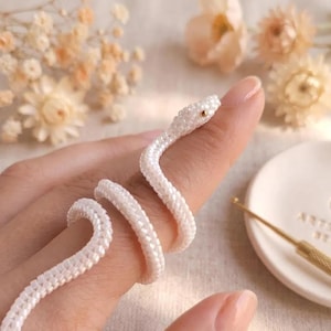 May include: A white beaded snake ring crafted from small, uniform seed beads. The snake features a single gold-colored bead for an eye and is coiled in a spiral shape. The design is flexible and wraps around the finger with a smooth, textured surface.