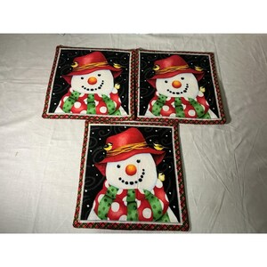 May include: Three square fabric pot holders featuring a snowman illustration on a black background. Each snowman wears a red hat with yellow birds and a green scarf with white polka dots. The borders are finished with a red and green plaid trim.