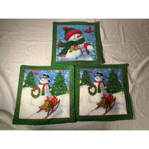 May include: Three square fabric potholders featuring a winter theme. One shows a snowman with a red hat and green scarf holding gifts. Two show a snowman with a black top hat and red scarf standing near a wooden sled with gifts and a wreath.