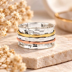 Puede incluir: Un anillo giratorio de varias bandas que presenta una banda base ancha de plata martillada, una banda central lisa de tono dorado y una banda lisa de tono oro rosa. El anillo está construido con tres bandas metálicas móviles apiladas.