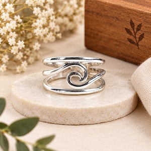 May include: A polished silver ring featuring a multi-band design that converges into a central wave-like spiral motif. The band is constructed from several smooth, interconnected silver wires that create an open, layered appearance on the finger.