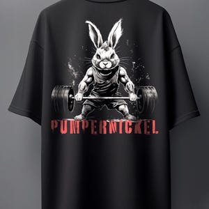 Könnte beinhalten: Ein schwarzes T-Shirt mit einer Grafik eines muskulösen Hasen, der eine Langhantel hebt. Der Hase trägt ein Unterhemd und eine kurze Hose. Unter dem Hasen ist das Wort PUMPERNICKEL in einer abgenutzten roten Schrift auf den Stoff gedruckt.