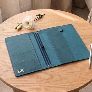 May include: A teal leather journal cover featuring three dark elastic bands for holding inserts, multiple interior pockets, and the gold embossed initials BK on the bottom left corner, accompanied by a silver metal pen.