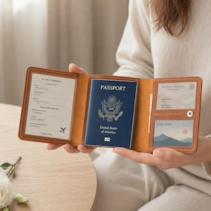 May include: A tan leather passport holder with visible stitching, featuring a clear pocket for emergency contact information, a central slot holding a blue United States passport, and two additional clear windows for membership and identification cards.
