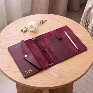 May include: A burgundy leather journal cover featuring a triangular pocket with a snap button, gold embossed initials MS, internal elastic bands for notebook inserts, a side pocket, and a silver pen clipped to the interior edge.