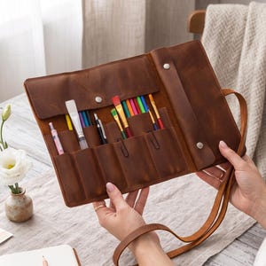 Könnte beinhalten: Eine braune Leder-Rolltasche mit mehreren Innenfächern, die mit verschiedenen bunten Pinseln, Stiften und Kugelschreibern gefüllt sind. Der Organizer verfügt über eine Klappe mit Metalldruckknopf und einen langen Lederriemen zum Tragen.