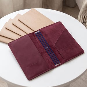 May include: A deep burgundy leather notebook cover featuring three vertical blue elastic bands for securing inserts, internal pockets on the right side, and a single horizontal slot on the left, accompanied by four tan paper notebooks.