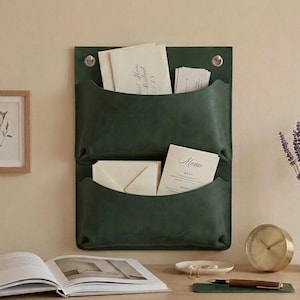 May include: A dark green wall-mounted organiser featuring two deep pockets for storing mail or documents. The organiser is constructed with a smooth finish and includes two silver-toned metal grommets at the top corners for secure wall attachment.