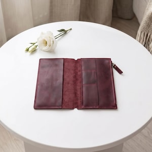 May include: A deep burgundy leather wallet laid open to reveal a smooth interior with multiple card slots and a zippered compartment on the right side. The wallet features a matte finish and visible stitching along the edges of the pockets.