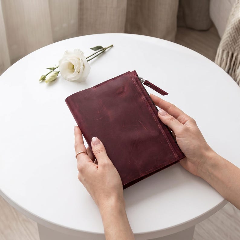 May include: A deep burgundy leather pouch featuring a smooth, matte finish with visible natural grain and texture. The rectangular accessory includes a side zipper closure with a matching leather pull tab, providing a secure opening for the main compartment.