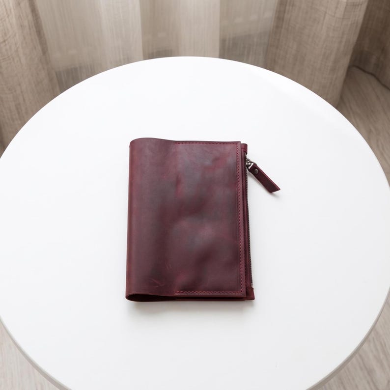 May include: A deep burgundy leather wallet featuring a smooth, matte finish and a vertical zippered compartment on the right side. The wallet has visible stitching along the edges and a matching leather zipper pull tab attached to the metal zipper.