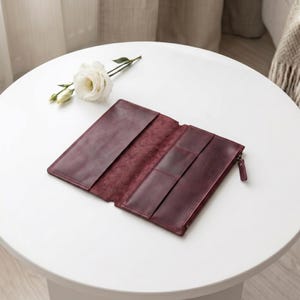 May include: A deep burgundy leather wallet laid open to reveal multiple interior card slots and a zippered compartment. The wallet features a smooth, matte finish and visible stitching along the edges, presented alongside a single white flower.
