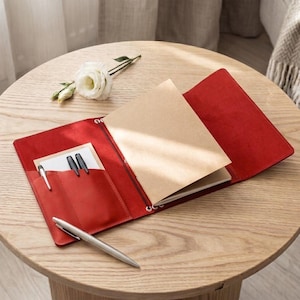 May include: A red leather notebook cover featuring an interior pocket with two black pens inserted, a silver pen resting on the surface, and a tan paper notebook secured by an elastic band. A white flower is positioned near the open notebook.