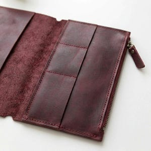 May include: A deep burgundy leather wallet featuring a smooth exterior finish and a textured interior surface. The wallet includes multiple vertical card slots and a side compartment secured with a metal zipper and a matching leather pull tab.