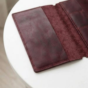 May include: A deep burgundy leather wallet or document holder featuring a smooth exterior panel and a textured interior section with visible stitching along the edges. The design includes multiple internal slots for organization and storage.