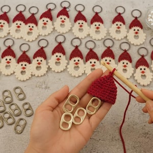 May include: A collection of crocheted Santa Claus keychains with red hats and white beards, featuring metal rings. A wooden crochet hook is actively working on a red yarn hat, with several loose metal soda can tabs nearby for assembly.