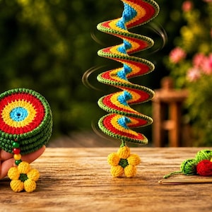 May include: A crocheted rainbow spiral wind spinner featuring a yellow flower at the bottom. Beside it, a stack of circular rainbow coasters with a green border and a yellow flower charm. Small yarn balls and a crochet hook are also present.