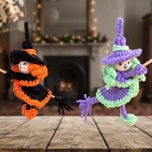 May include: Two crocheted witch ornaments hanging in the air. One witch wears a black and orange outfit with a matching hat, while the other wears a purple and light green outfit with a matching hat. Both figures hold a small wooden broomstick.