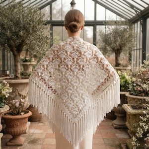May include: A triangular off-white crochet shawl featuring an intricate floral pattern and long fringe along the bottom edges. The shawl is draped over the shoulders, showcasing the detailed needlework and the delicate, open-weave texture.