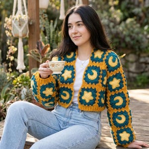 May include: A cropped cardigan made of crocheted granny squares in mustard yellow and dark teal. The squares feature alternating yellow star and crescent moon motifs. The garment has long sleeves and an open front, worn over a plain white t-shirt.