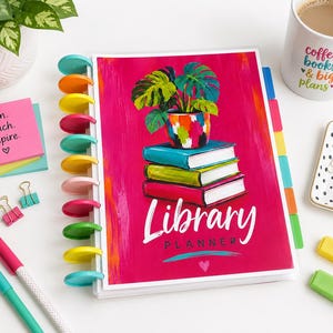 May include: A disc-bound Library Planner with a vibrant magenta cover featuring a stack of three books topped by a potted plant. The cover displays the text Library Planner in white script. The planner has colourful plastic discs along the spine.