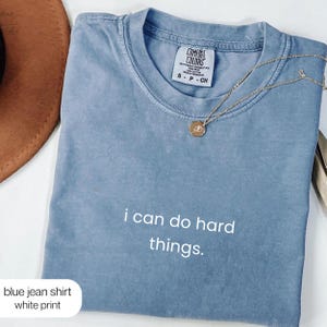 May include: A blue short-sleeved t-shirt with a crew neckline and a Comfort Colors label at the collar. The shirt features the white text "i can do hard things." printed on the front. A gold-toned chain necklace with a small round pendant is visible.