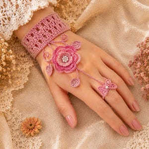 Puede incluir: Una pulsera de mano tejida a ganchillo en color rosa empolvado que presenta una banda ancha con patrón geométrico, un motivo central de rosa en capas con centro de perlas, detalles de hojas delicadas y un anillo de mariposa a juego.