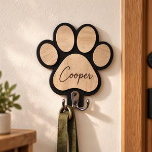 Può includere: Un gancio da parete a forma di impronta di zampa con cornice nera e inserti in legno chiaro. Il cuscinetto centrale presenta il nome Cooper inciso in corsivo. Un gancio in metallo argentato fissato sul fondo sostiene un guinzaglio in tessuto verde.