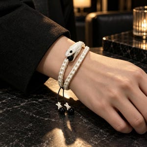 Può includere: Un bracciale avvolgente con una testa di serpente bianca intagliata a un'estremità e una serie di segmenti bianchi simili a ossa che formano il cinturino. Il design include una piccola perla nera nella bocca del serpente e due perle alle estremità.