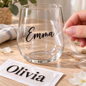 Könnte beinhalten: Ein klares Glas mit dem in schwarzer Schreibschrift aufgebrachten Namen Emma, wobei eine transparente Transferfolie abgezogen wird. Ein separates rechteckiges weißes Papier zeigt den in schwarzer Serifenschrift gedruckten Namen Olivia.