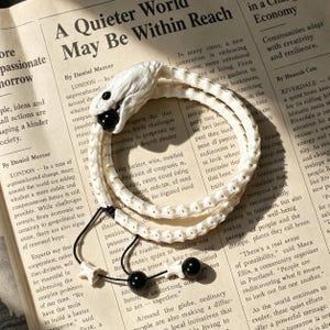 Può includere: Un bracciale a forma di serpente avvolto con un corpo testurizzato bianco sporco e una testa dettagliata che tiene una perla nera liscia in bocca. Il bracciale include cordini neri con due perle nere aggiuntive e piccoli dettagli a forma di osso.