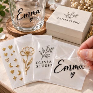 Puede incluir: Un vaso de cristal transparente con el nombre Emma y un corazón impresos en negro, una caja de regalo blanca con la etiqueta Olivia Studio y varias hojas de pegatinas con corazones, estrellas, lunas doradas, una flor y el nombre Emma.