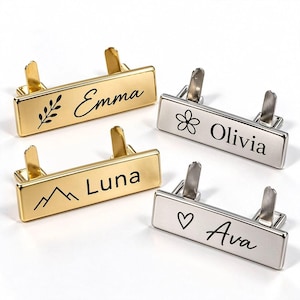 May include: Four rectangular metal name tags with two prongs on the back. Two are gold-toned and two are silver-toned. They feature black engraved names and icons: Emma with a leaf, Olivia with a flower, Luna with mountains, and Ava with a heart.