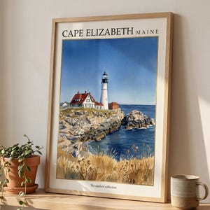 May include: A framed watercolor print featuring a lighthouse on a rocky coastline in Cape Elizabeth, Maine. The artwork depicts a white lighthouse, red-roofed buildings, blue ocean water, and golden grasses. The text reads CAPE ELIZABETH MAINE and The outdoor collection.