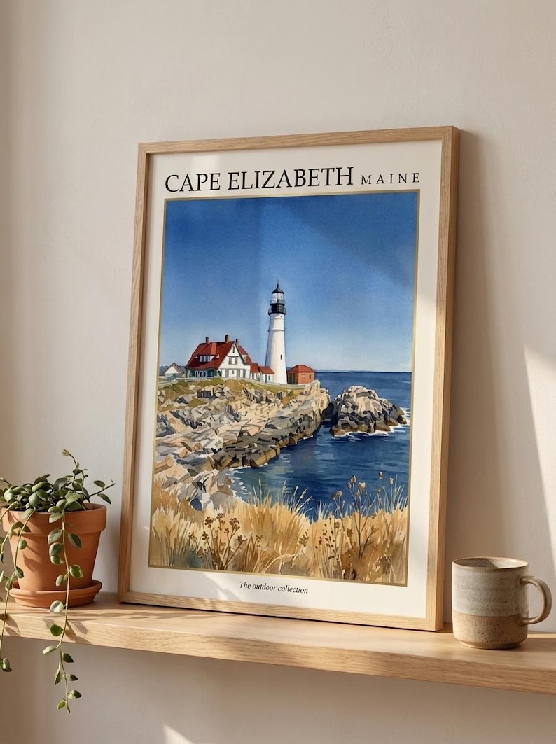 May include: A framed watercolor print featuring a lighthouse on a rocky coastline in Cape Elizabeth, Maine. The artwork depicts a white lighthouse, red-roofed buildings, blue ocean water, and golden grasses. The text reads CAPE ELIZABETH MAINE and The outdoor collection.