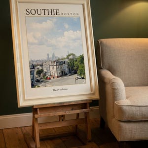 May include: A framed print titled SOUTHIE BOSTON featuring a cityscape view of residential buildings and a distant skyline under a blue sky with clouds. The text The city collection is printed at the bottom of the white matting within the frame.