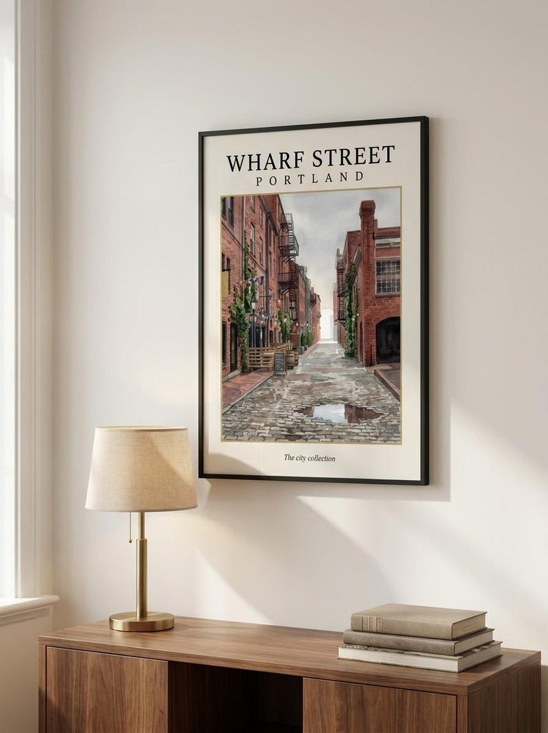 May include: A framed vertical wall art print titled WHARF STREET PORTLAND. The image depicts a cobblestone street lined with red brick buildings, fire escapes, and green vines. A puddle reflects the sky on the ground. The text The city collection appears at the bottom.