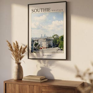 May include: A framed wall art print featuring a photograph of a South Boston neighborhood street scene under a blue sky with clouds. The text SOUTHIE BOSTON is printed at the top, and The city collection is printed at the bottom of the print.