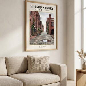May include: A framed wall art print titled WHARF STREET PORTLAND from The city collection. The print features a perspective view of a cobblestone street lined with red brick buildings, green climbing vines, and a small puddle in the center.