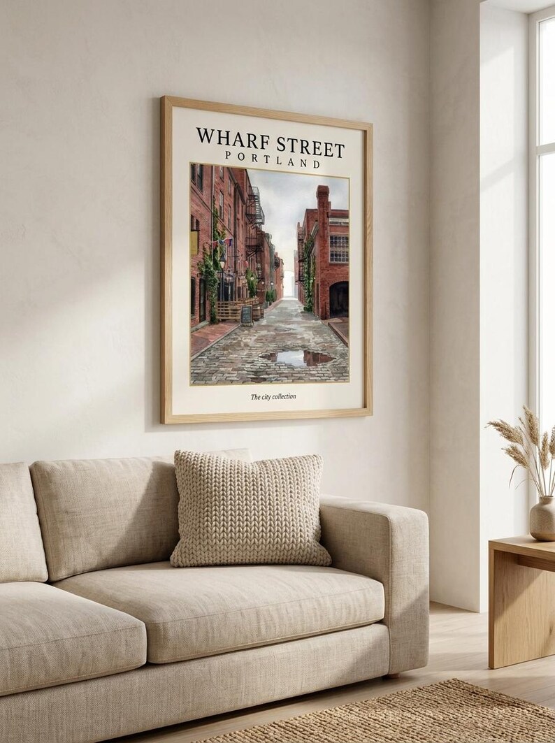 May include: A framed wall art print titled WHARF STREET PORTLAND from The city collection. The print features a perspective view of a cobblestone street lined with red brick buildings, green climbing vines, and a small puddle in the center.