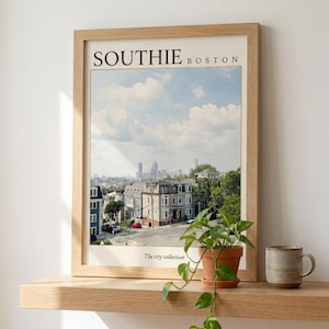 May include: A framed wall art print titled SOUTHIE BOSTON with the text The city collection at the bottom. The print features a daytime cityscape view of Boston with residential buildings in the foreground and a skyline under a blue sky with clouds.