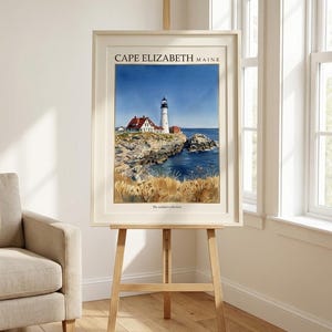 May include: A framed art print featuring a watercolor painting of a lighthouse on a rocky coastline in Cape Elizabeth, Maine. The scene includes a white lighthouse, red-roofed buildings, blue water, and golden grasses. The text reads CAPE ELIZABETH MAINE.