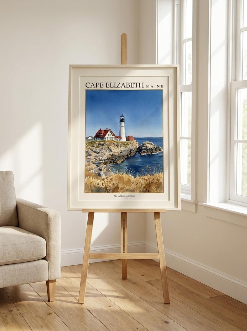 May include: A framed art print featuring a watercolor painting of a lighthouse on a rocky coastline in Cape Elizabeth, Maine. The scene includes a white lighthouse, red-roofed buildings, blue water, and golden grasses. The text reads CAPE ELIZABETH MAINE.