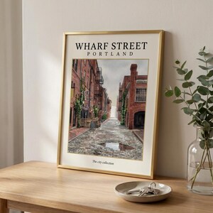 May include: A framed art print featuring a watercolor illustration of Wharf Street in Portland, with red brick buildings, a cobblestone road, and a puddle. The text reads WHARF STREET PORTLAND and The city collection in a gold metal frame.