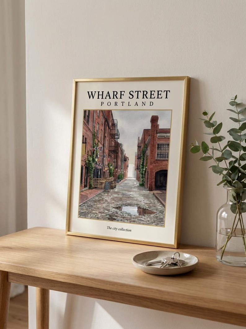 May include: A framed art print featuring a watercolor illustration of Wharf Street in Portland, with red brick buildings, a cobblestone road, and a puddle. The text reads WHARF STREET PORTLAND and The city collection in a gold metal frame.