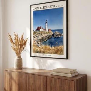 May include: A framed wall art print featuring a watercolor illustration of the Cape Elizabeth Maine lighthouse. The scene depicts a white lighthouse, a red-roofed building, rocky cliffs, blue ocean water, and golden grasses. The text reads CAPE ELIZABETH MAINE.