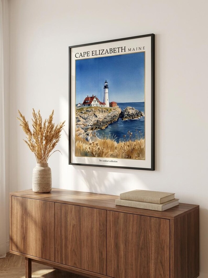 May include: A framed wall art print featuring a watercolor illustration of the Cape Elizabeth Maine lighthouse. The scene depicts a white lighthouse, a red-roofed building, rocky cliffs, blue ocean water, and golden grasses. The text reads CAPE ELIZABETH MAINE.