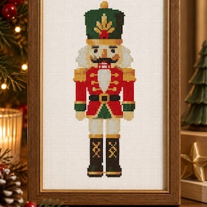 May include: A framed cross-stitch embroidery featuring a nutcracker soldier. The figure wears a green hat with a gold emblem, a red jacket with gold trim, a white beard, and black boots. The design is stitched on white Aida cloth in a wooden frame.
