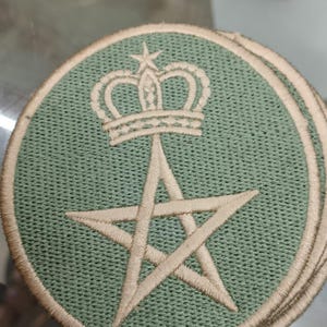May include: A circular embroidered patch featuring a sage green background with a tan border. The design includes a tan five-pointed star centered below a tan crown. The stitching is visible throughout the fabric, creating a textured appearance.